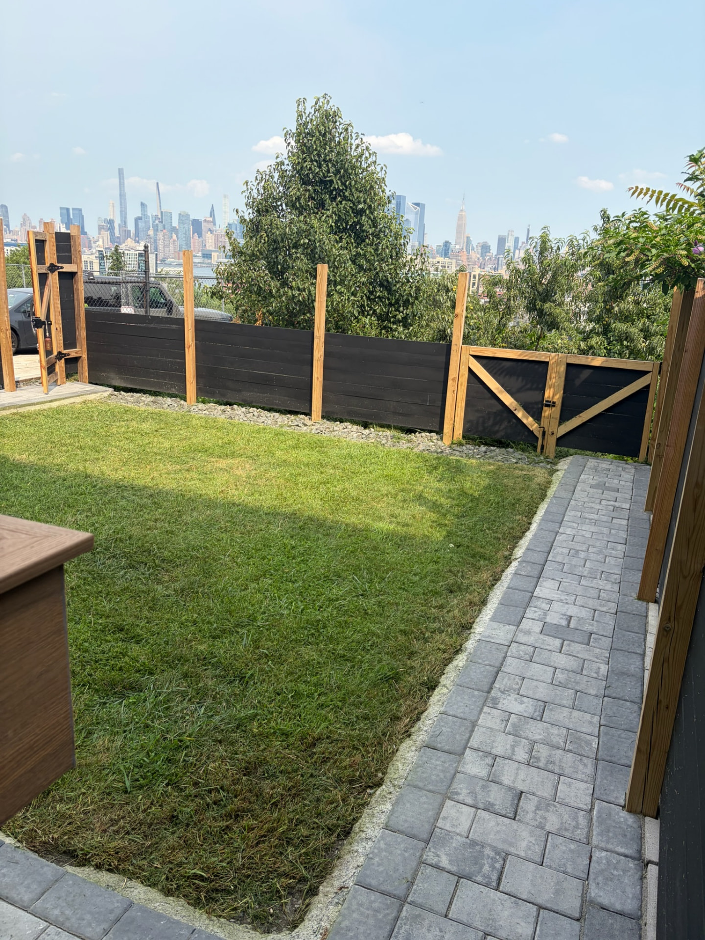 Green turf and stone paver path beside a wood fence and gate, with a city skyline behind.